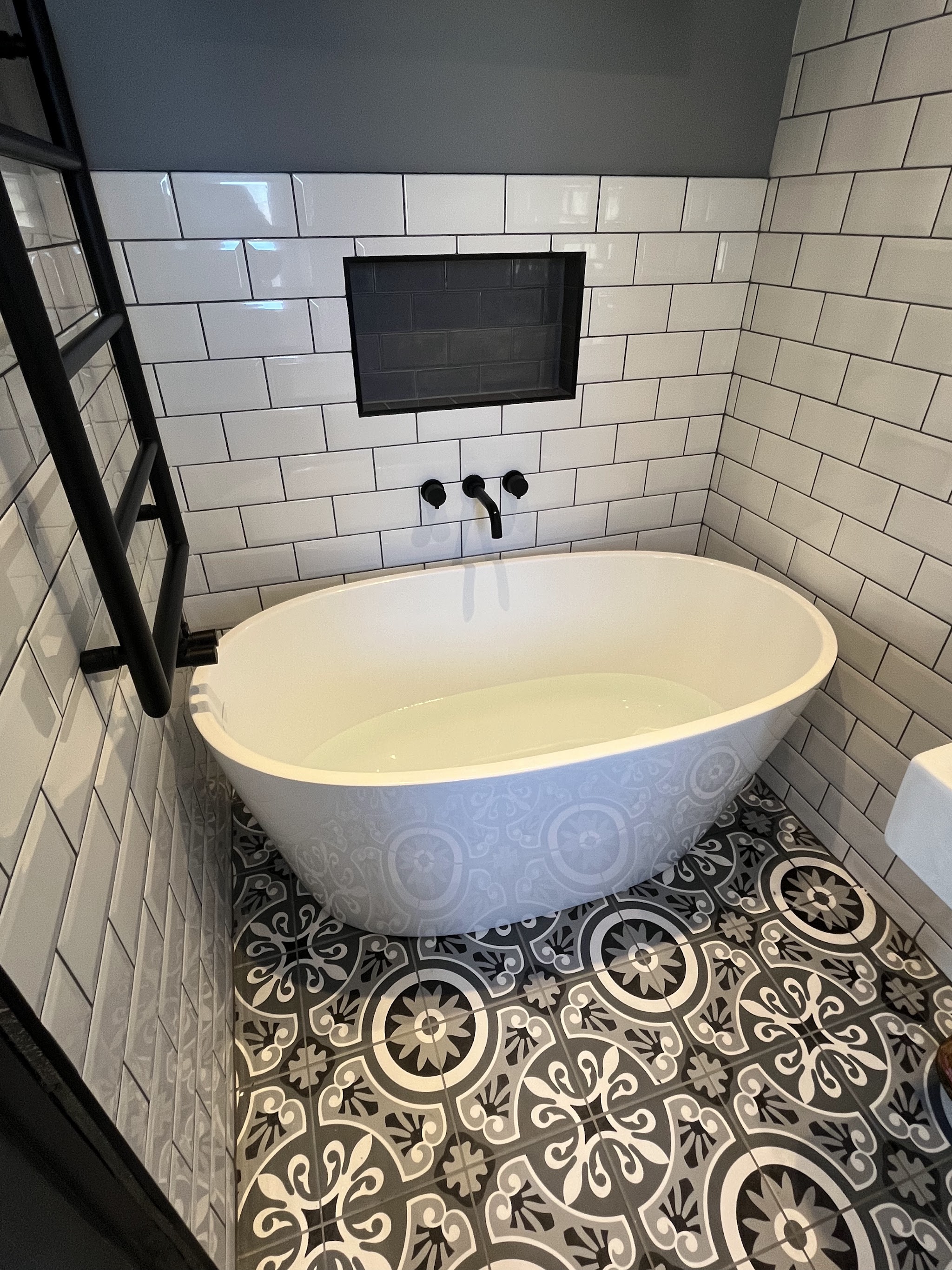 Freestanding bath with matt black wall-mounted taps, a tiled niche and patterned black and white floor tiles